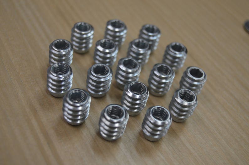 May include: A close-up of a group of silver metal threaded inserts. The inserts are arranged in a circular pattern on a wooden surface.