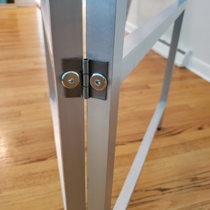 May include: Close-up of a silver metal frame with a hinge. The hinge has two silver screws.