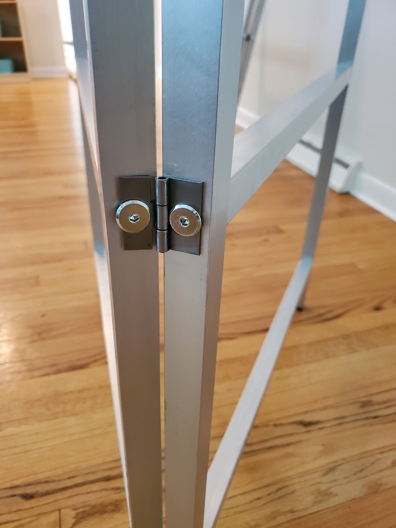 May include: Close-up of a silver metal frame with a hinge. The hinge has two silver screws.