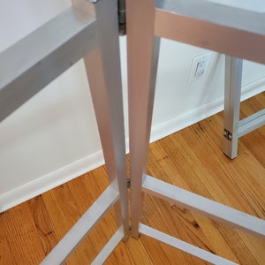 May include: A silver metal folding table frame with a hinge in the center. The frame is made of square tubing and has a simple design.