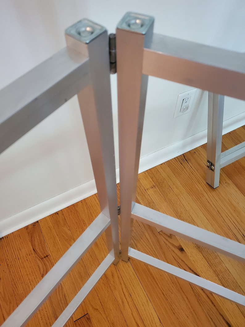 May include: A silver metal folding table frame with a hinge in the center. The frame is made of square tubing and has a simple design.