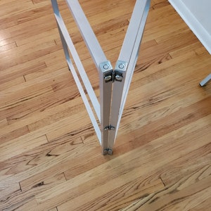 May include: Close-up of a silver metal folding table frame with two hinges. The frame is on a wooden floor.