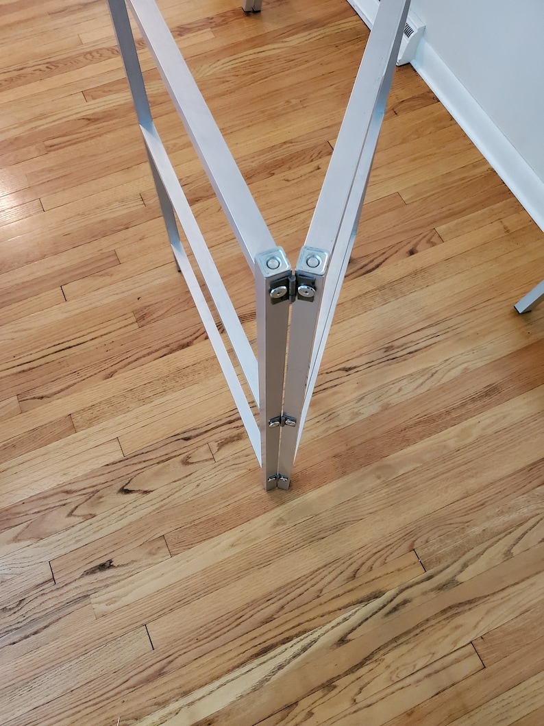 May include: Close-up of a silver metal folding table frame with two hinges. The frame is on a wooden floor.