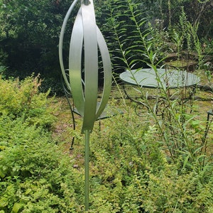 May include: A silver metal sculpture of a swirling leaf design, standing tall in a garden setting. The sculpture is made of multiple curved metal pieces that create a dynamic and abstract form.