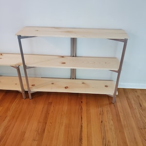 May include: A three-tiered wooden shelf unit with silver metal supports. The shelves are made of unfinished wood and the metal supports are a light silver color. The shelf unit is standing on a hardwood floor.