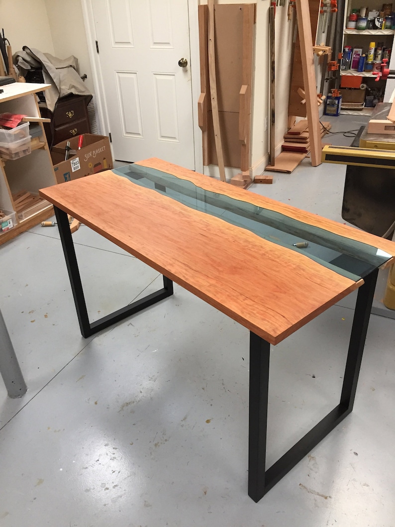 May include: A wooden table with a black metal frame. The table top is made of wood with a clear epoxy river running through the center. The table is in a workshop setting with tools and other wood pieces in the background.