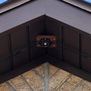 Puede incluir: Esquina exterior de un edificio con techo de madera marrón y fachada de piedra y ladrillo. Pequeñas luces redondas están montadas en la parte inferior del techo, conectadas por cables negros. El cielo es visible en el fondo.