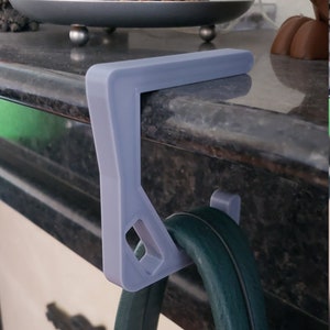 May include: A light gray, 3D-printed purse hanger designed to hook over a shelf or ledge. The hanger is supporting a dark green purse. The design includes a rectangular top and a hook for the purse strap.