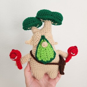 May include: A handmade crocheted tree-like figure with a tan body, green leafy head, and red accessories. The figure has a green leaf-shaped detail on its chest and is holding two red objects. The figure is made with a crochet technique.