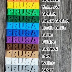 May include: A vertical display of Prusa 3D printer filament samples in various colors, including pink, red, orange, yellow, green, blue, purple, brown, tan, white, gray, black, silver, and gold. Each sample has the word "PRUSA" printed on it.