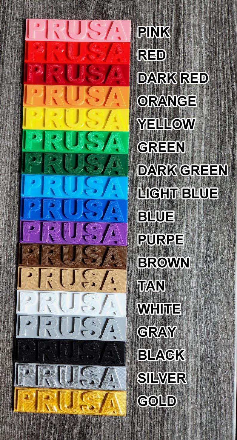 May include: A vertical display of Prusa 3D printer filament samples in various colors, including pink, red, orange, yellow, green, blue, purple, brown, tan, white, gray, black, silver, and gold. Each sample has the word "PRUSA" printed on it.