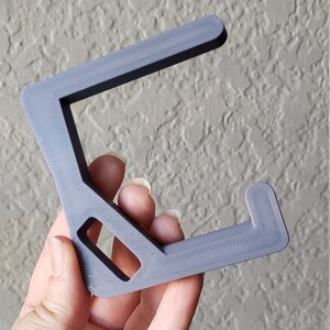 May include: A gray, 3D-printed headphone stand. The stand has a C-shape with a rectangular opening and a small hook at the end. The stand is held by a hand against a textured wall.