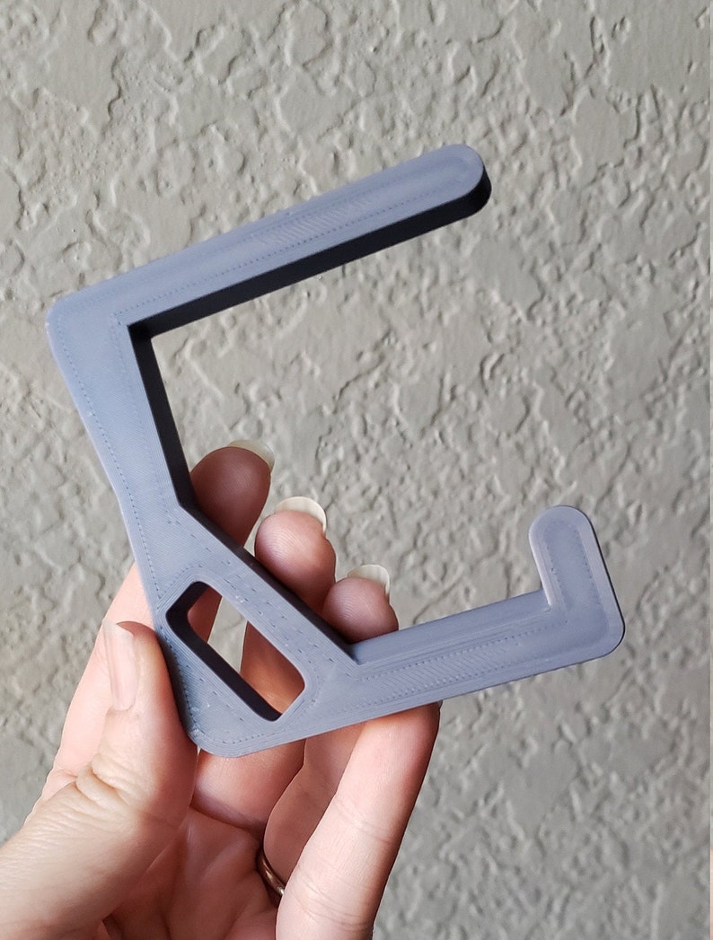 May include: A gray, 3D-printed headphone stand. The stand has a C-shape with a rectangular opening and a small hook at the end. The stand is held by a hand against a textured wall.