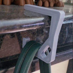 May include: A light gray purse hook with a diamond-shaped cutout, designed to hang a green handbag. The hook is attached to a surface, with two brown owl figurines in the background.
