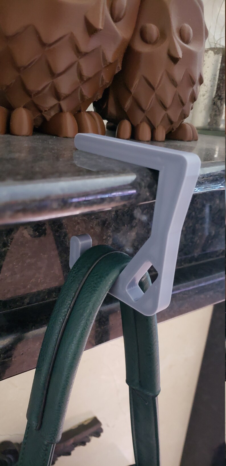 May include: A light gray purse hook with a diamond-shaped cutout, designed to hang a green handbag. The hook is attached to a surface, with two brown owl figurines in the background.