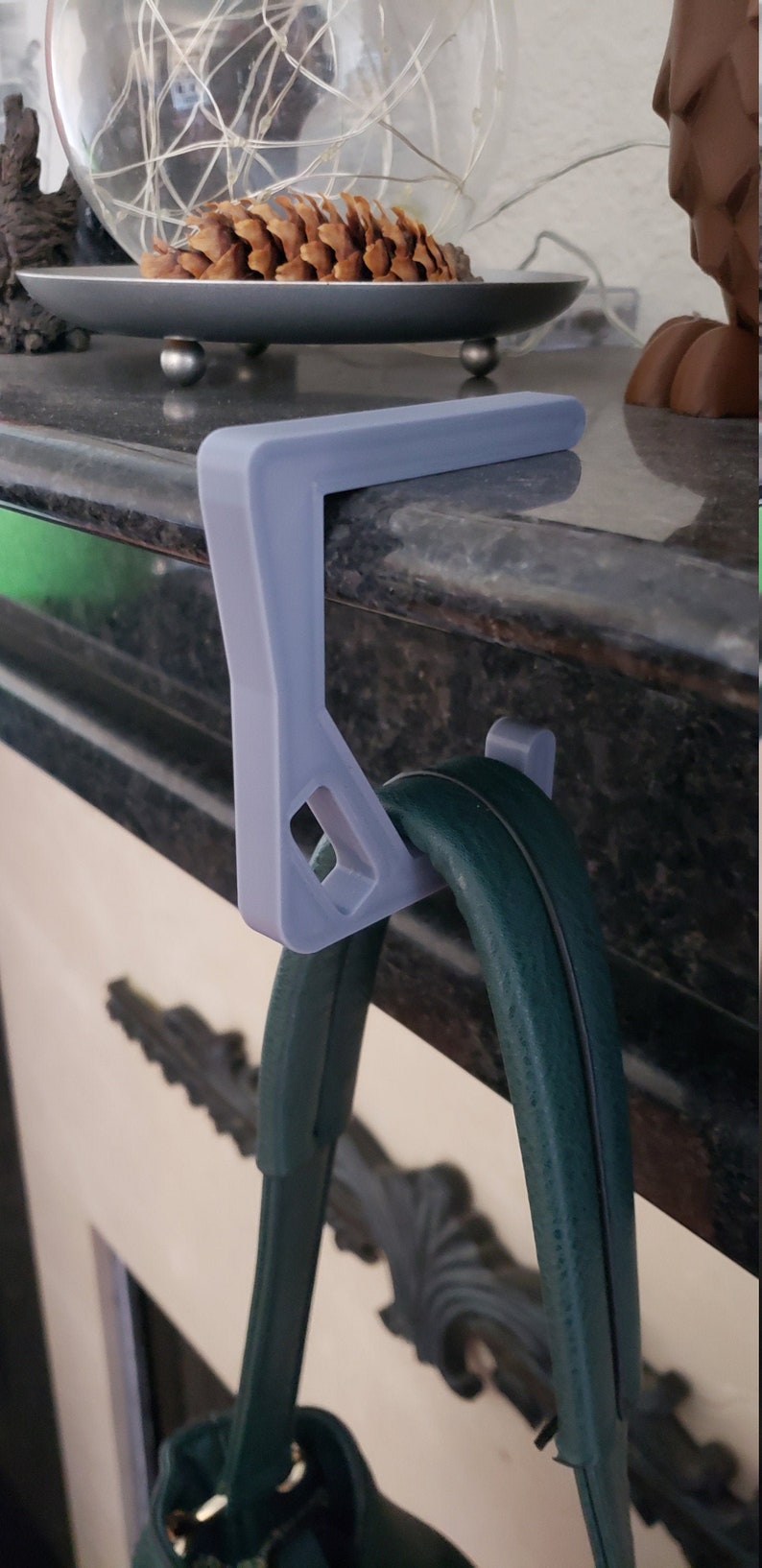 May include: A light gray, 3D-printed purse hanger designed to hook over a shelf or ledge. The hanger is supporting a dark green purse. The design includes a rectangular top and a hook for the purse strap.