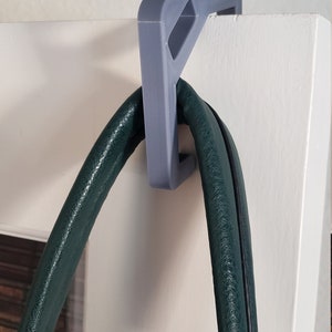 May include: A gray, 3D-printed door hanger designed to hold a purse or bag. The hanger is mounted on a white door and supports a dark green leather bag.