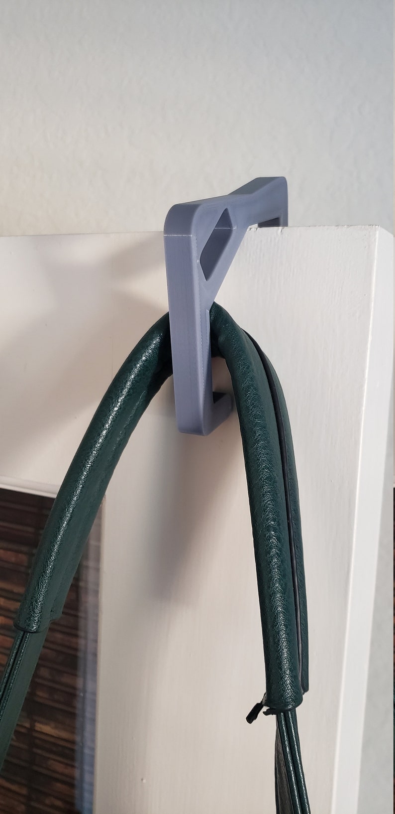 May include: A gray, 3D-printed door hanger designed to hold a purse or bag. The hanger is mounted on a white door and supports a dark green leather bag.