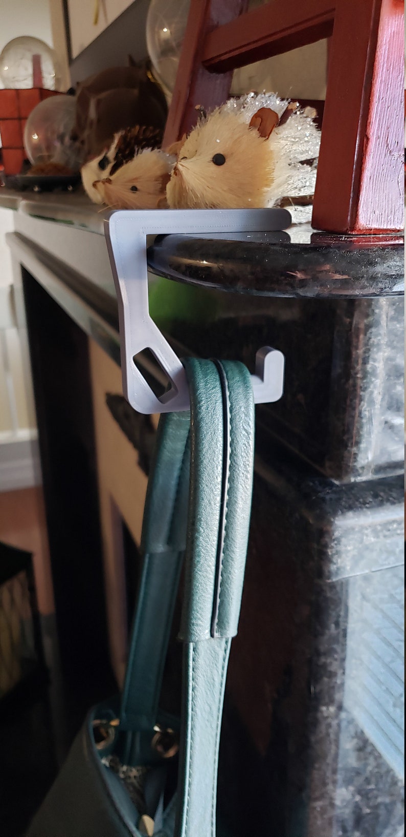 May include: A gray, 3D-printed hook designed to hang a purse. The hook is attached to a dark surface, and a teal purse hangs from it. Decorative hedgehog figurines are visible in the background.