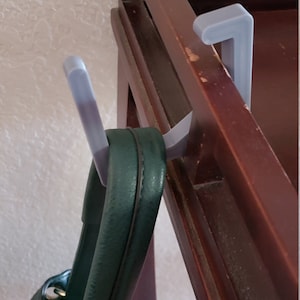 May include: A dark brown wooden coat rack with a green handbag hanging from a gray plastic hook. The hook is attached to the rack. The handbag has a leopard print lining.
