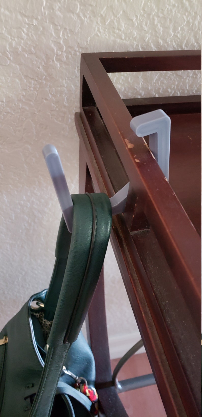 May include: A dark brown wooden coat rack with a green handbag hanging from a gray plastic hook. The hook is attached to the rack. The handbag has a leopard print lining.