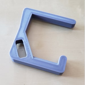 May include: A lavender-colored, 3D-printed object with a unique, angular design. It features a rectangular opening and a curved arm, likely a tool or accessory. The object is resting on a light-colored wooden surface.