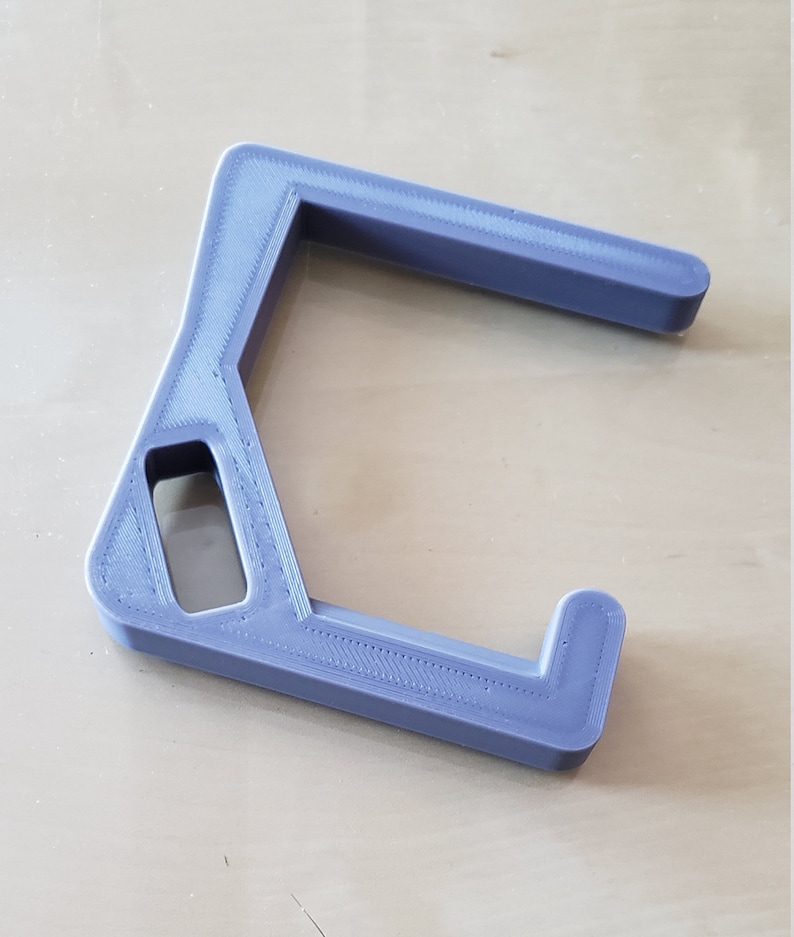 May include: A lavender-colored, 3D-printed object with a unique, angular design. It features a rectangular opening and a curved arm, likely a tool or accessory. The object is resting on a light-colored wooden surface.