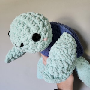 May include: A crocheted plush toy turtle with a light blue and purple shell. The turtle has black eyes and a pink nose.