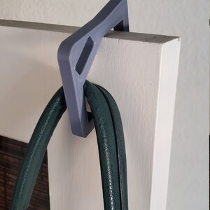 May include: A gray over-the-door hook holds a dark green handbag. The hook is made of a solid material and is designed to fit over a door. The handbag has a textured surface.