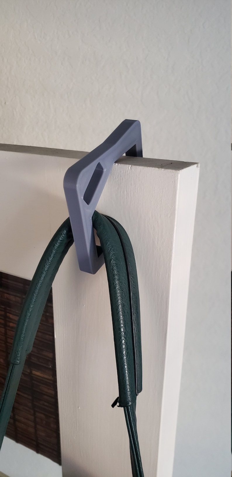 May include: A gray over-the-door hook holds a dark green handbag. The hook is made of a solid material and is designed to fit over a door. The handbag has a textured surface.