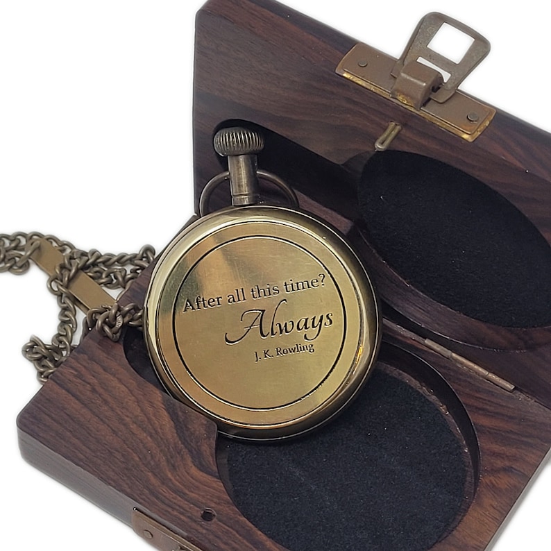 May include: A brass pocket watch with the inscription "After all this time? Always. J.K. Rowling" is displayed open in a dark wooden box. The watch has a chain and is set against a black velvet interior.