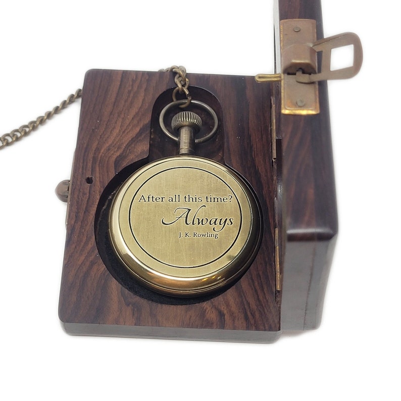 May include: A brass pocket watch engraved with "After all this time? Always. J.K. Rowling" sits in an open dark wooden box. The box has a brass clasp and a chain attached to the watch.