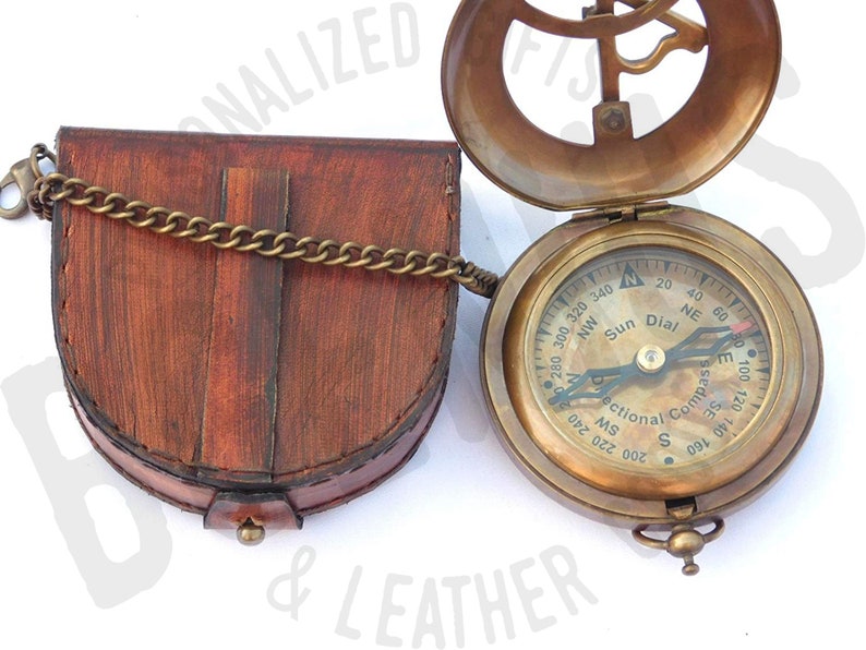 May include: A brass compass with a sun dial and a leather case. The compass has a black face with white markings and a black needle. The case is brown and has a chain attached to it.