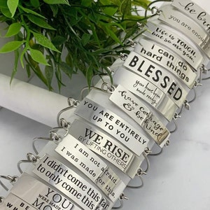 May include: A stack of silver bracelets with inspirational messages engraved on each. The messages include "be brave", "life is tough", "i can do hard things", "blessed", "have courage & be kind", "you are entirely up to you", "we rise by lifting others", "i am not afraid", "i didn't come this far to only come this far", and "you were made more".
