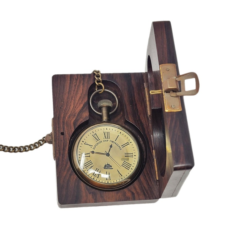 May include: A vintage pocket watch with a gold-tone face and Roman numerals, presented in a dark wood case. The watch is attached to a chain and the case has a brass clasp. The watch face reads "SUPERIOR SHIP TIMEKEEPER".