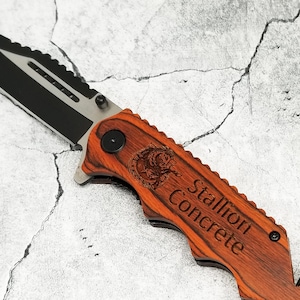 May include: A brown wooden folding pocket knife with a black blade and a safety feature. The handle is engraved with the words "Stallion Concrete" and a stylized image of a horse.