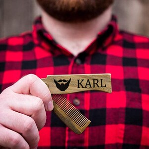 May include: A wooden beard comb with a personalized name "KARL" and a beard icon engraved on the front. The comb is closed and held in a hand.