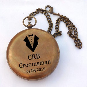 May include: A brass compass with a chain, engraved with a tuxedo graphic, the initials "CRB", the word "Groomsman", and the date "6/29/2019".