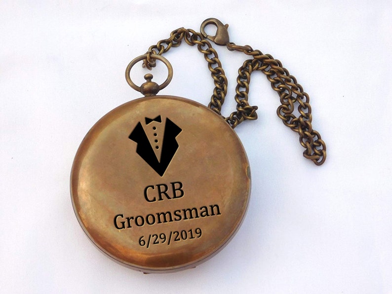 May include: A brass compass with a chain, engraved with a tuxedo graphic, the initials "CRB", the word "Groomsman", and the date "6/29/2019".