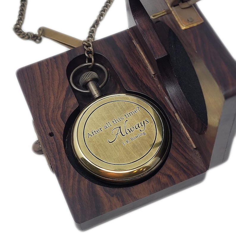May include: A brass pocket watch engraved with "After all this time? Always" by J.K. Rowling, presented in an open wooden box. The watch is suspended by a chain and set against a dark interior.