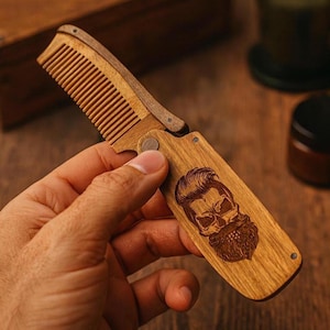 May include: A wooden folding comb with a skull design etched on the handle. The comb is made of light brown wood and has fine teeth. The handle features a detailed illustration of a skull with a beard.