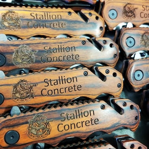 May include: Close-up of a stack of wooden pocket knives with a black metal blade and a black button. Each knife has a brown and black wood grain pattern and is engraved with the text "Stallion Concrete".