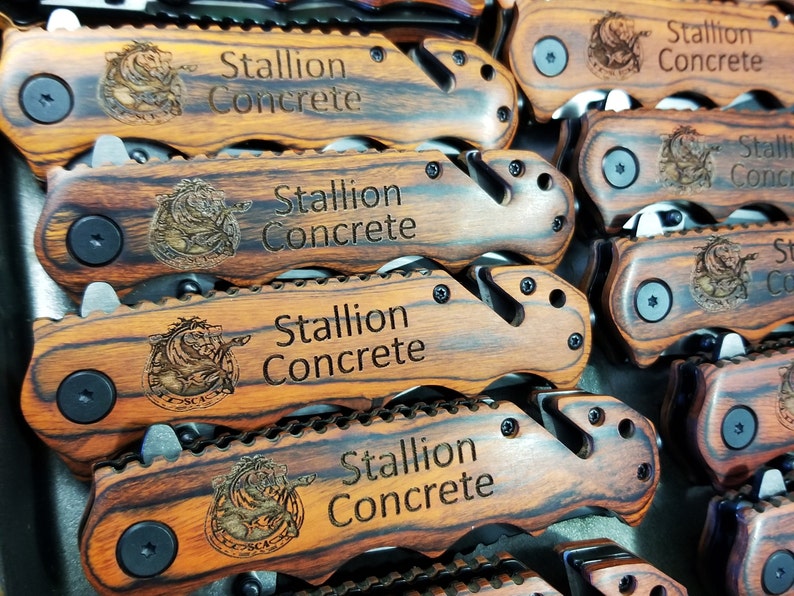 May include: Close-up of a stack of wooden pocket knives with a black metal blade and a black button. Each knife has a brown and black wood grain pattern and is engraved with the text "Stallion Concrete".
