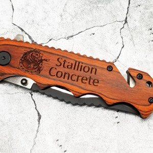 May include: A brown wooden folding knife with a black blade and a safety feature. The knife has a black metal button and a silver metal rivet. The handle is engraved with the words "Stallion Concrete" and a stylized image of a horse's head.