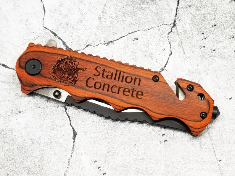 May include: A brown wooden folding knife with a black blade and a safety feature. The knife has a black metal button and a silver metal rivet. The handle is engraved with the words "Stallion Concrete" and a stylized image of a horse's head.