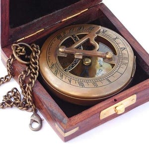May include: A brass compass with a wooden box and a chain. The compass has a sun dial and a compass rose. The box is closed.