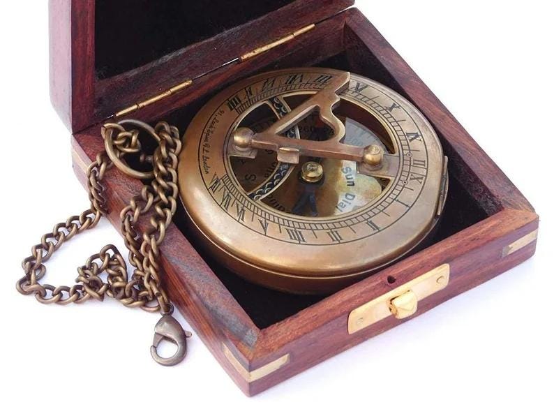 May include: A brass compass with a wooden box and a chain. The compass has a sun dial and a compass rose. The box is closed.