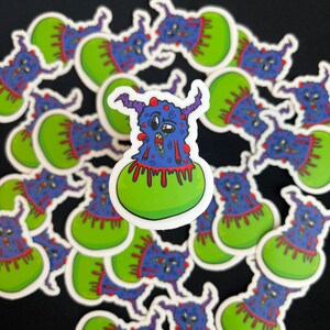 Op de afbeelding: Een verzameling stickers met een blauw en paars monster met rode, druipende slijm op een groene achtergrond. Het monster heeft een verraste uitdrukking met grote ogen en een brede mond.