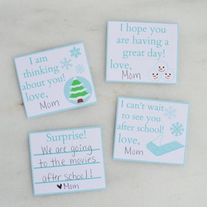 Op de afbeelding: Vier blauwe en witte kaarten met winterse illustraties en handgeschreven berichten. De berichten zijn: "Ik denk aan je! Liefs, Mama", "Verrassing! We gaan na school naar de bioscoop! Liefs, Mama", "Ik hoop dat je een fijne dag hebt! Liefs, Mama", en "Ik kan niet wachten om je na school te zien! Liefs, Mama".