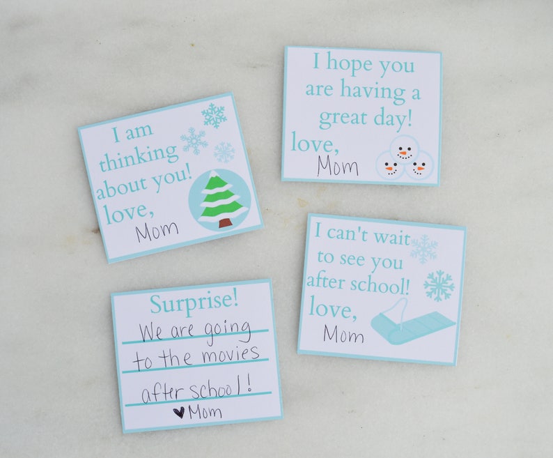 Op de afbeelding: Vier blauwe en witte kaarten met winterse illustraties en handgeschreven berichten. De berichten zijn: "Ik denk aan je! Liefs, Mama", "Verrassing! We gaan na school naar de bioscoop! Liefs, Mama", "Ik hoop dat je een fijne dag hebt! Liefs, Mama", en "Ik kan niet wachten om je na school te zien! Liefs, Mama".